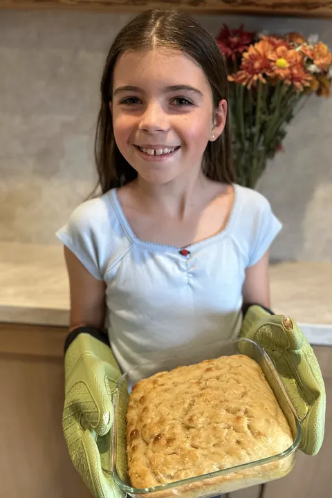 Ruby smiling in the kitchen, holding a fresh pan of focaccia in green oven mitts.